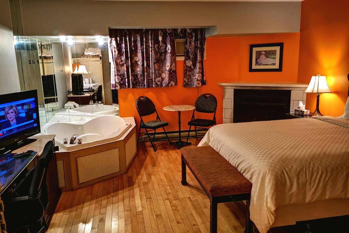 A hotel room with a large bed, a sitting area with a table and chairs, a bathtub, a television, and a fireplace.
