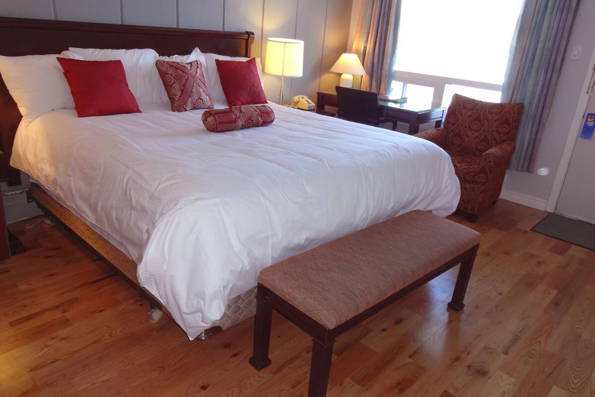 A neatly made bed with white and red bedding, a wooden bench, and a chair in a hotel room with wooden flooring and a window with curtains.