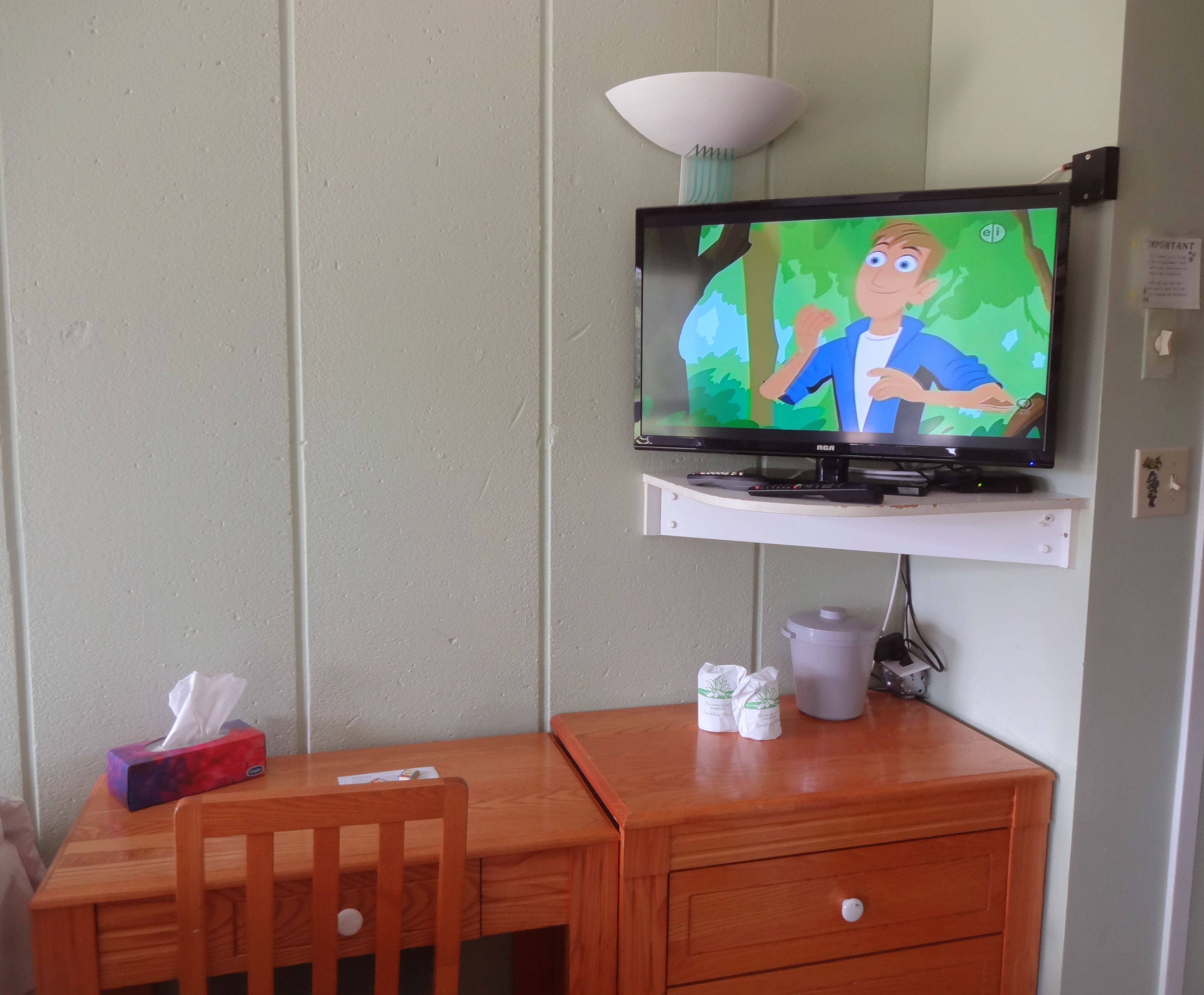 A television mounted on a wall above a wooden desk with a chair, a tissue box, and a small trash can.