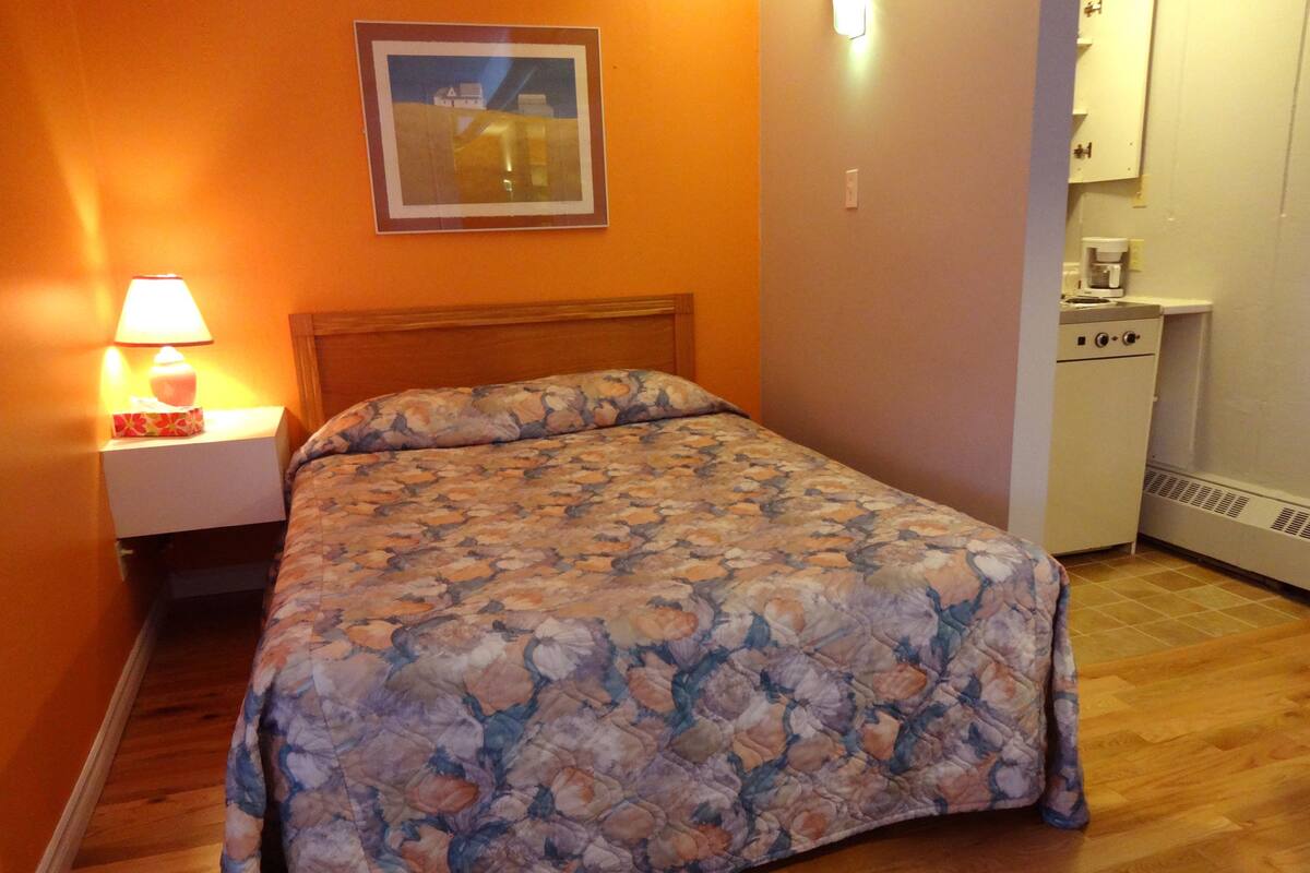 A bedroom with a floral bedspread, a nightstand with a lamp, and a small kitchenette area.