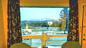A room with two green chairs, a small table with a lamp, and a view of a snowy landscape with mountains. - Motel Tremblant (Mont-Blanc)