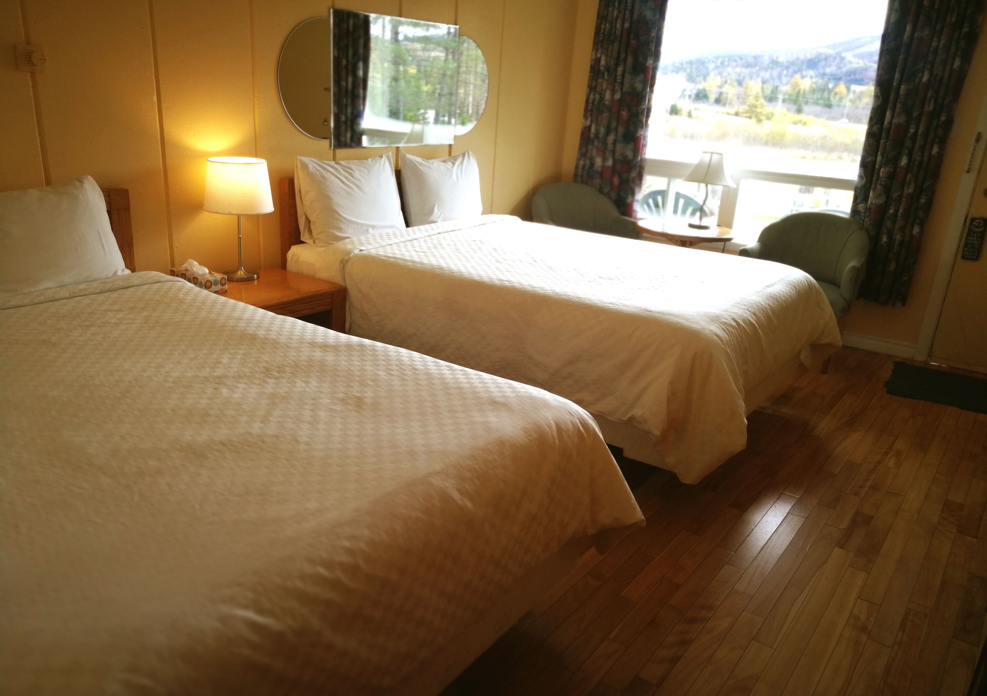 A hotel room with two beds, a nightstand with a lamp, a chair, and a view of the outdoors.
