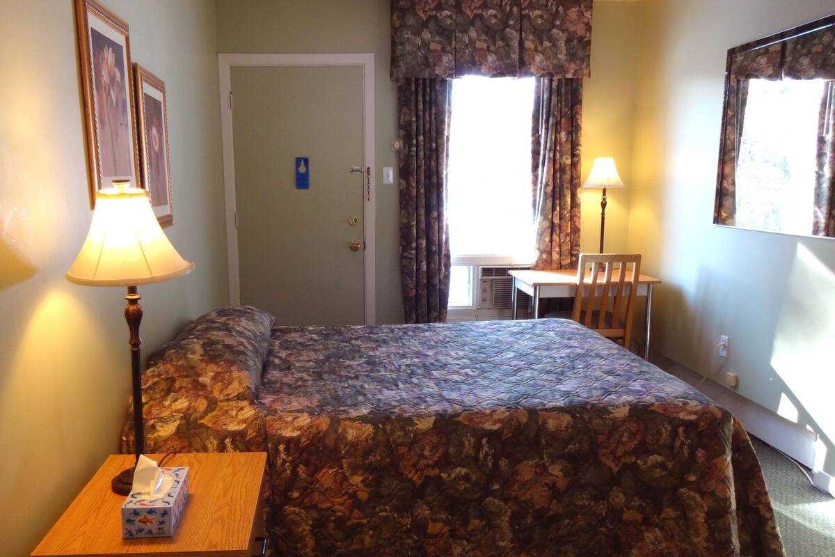 A hotel room with a bed, a desk, a chair, a lamp, and a window with curtains.