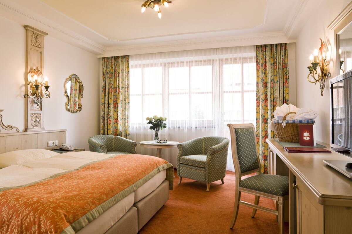 A hotel room with a bed, two armchairs, a desk with a laptop, a television, and a window with floral curtains.