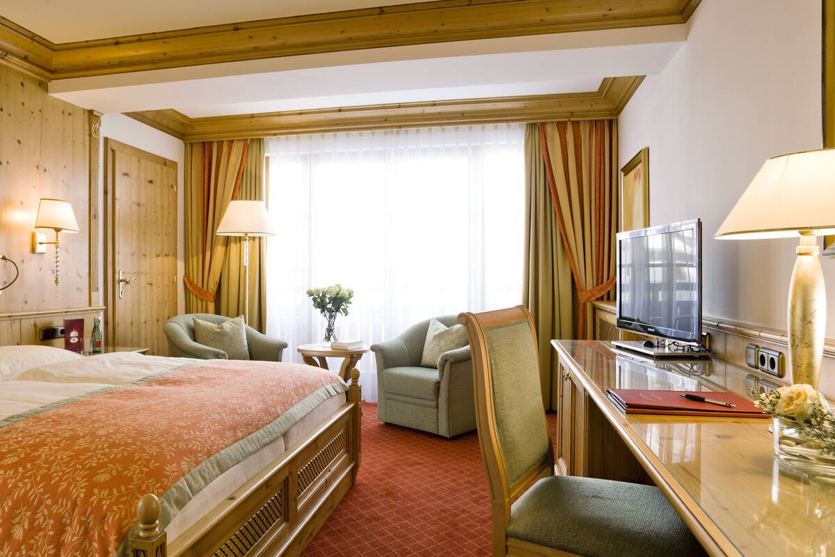 A hotel room with a large bed, two armchairs, a desk with a TV, and a window with curtains.