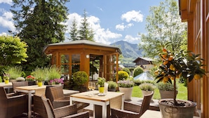 An outdoor seating area with wicker chairs and tables, surrounded by potted plants and a wooden gazebo.
