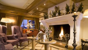 A cozy living room with a fireplace, plush armchairs, a coffee table, and a view of the city at dusk.