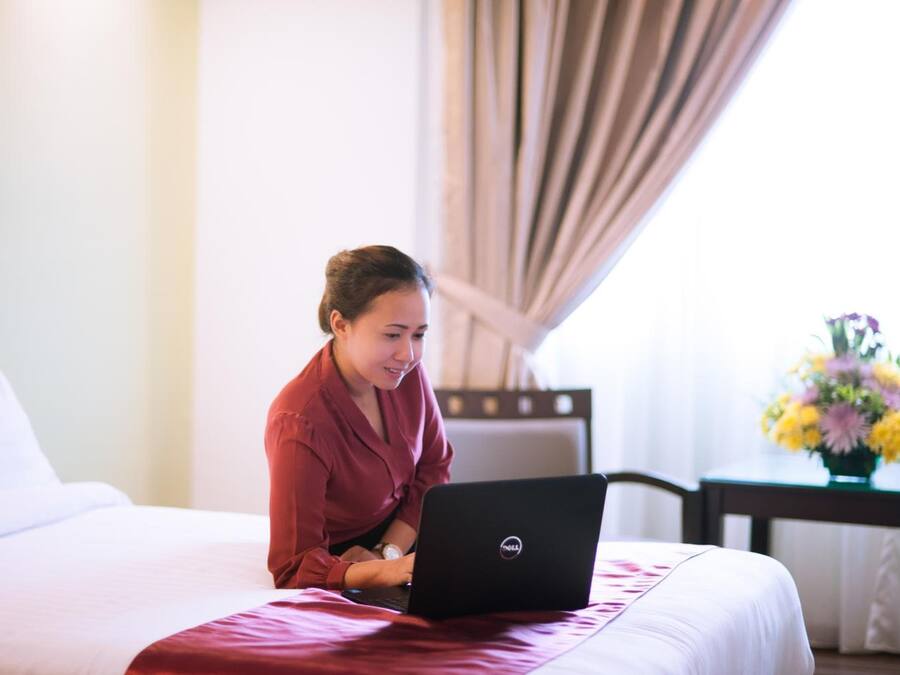 A person sitting on a bed with a laptop, a vase of flowers, and a window with curtains.