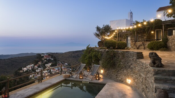 Una piscina con sillones de descanso y un muro de piedra, con vistas a un pueblo costero con casas e iglesia.