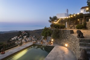 Une piscine bordée de chaises longues et adossée à un mur de pierre, d’où l’on a vue sur un village côtier avec ses maisons et son église.