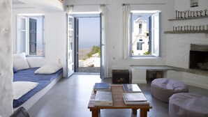 Una habitación con cama, una mesa con libros, chimenea y vistas al mar a través de puertas abiertas.