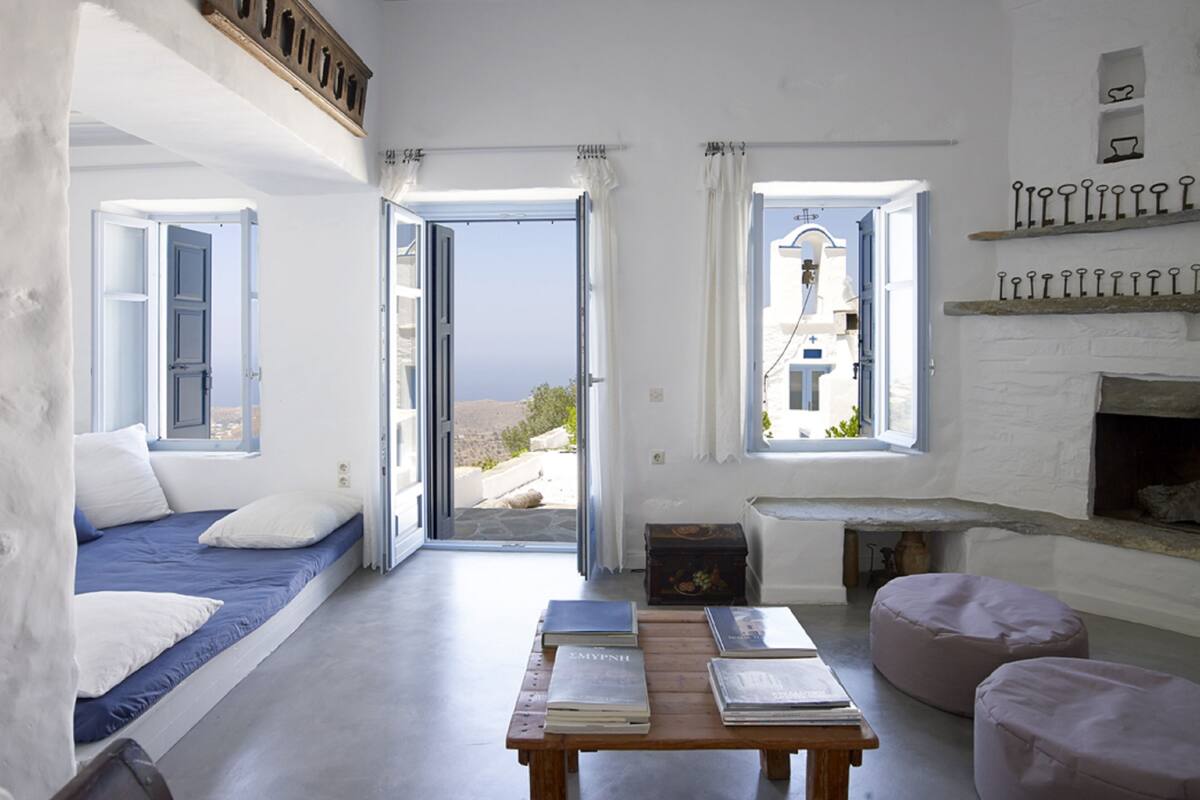 Ein Zimmer mit einem Bett, einem Tisch mit Büchern, einem Kamin und Meerblick durch offene Türen.
