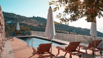 Eine Holzterrasse mit Liegestühlen und Sonnenschirmen mit Blick auf einen Pool, mit Hügeln im Hintergrund.