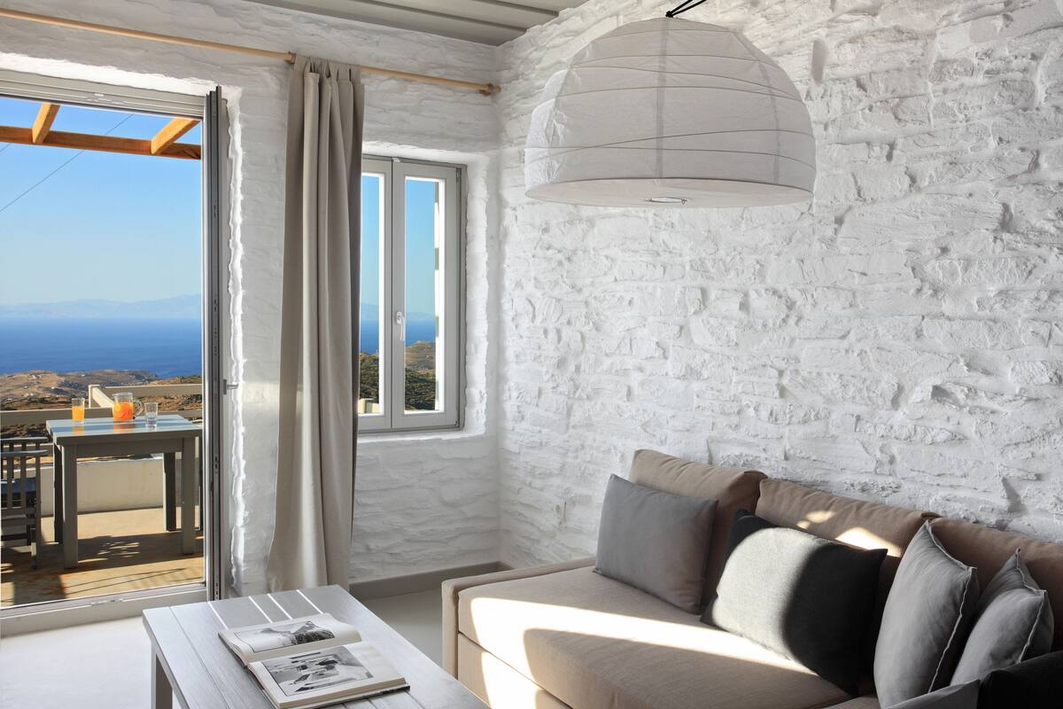 Ein gemütliches Zimmer mit Steinwand, einer Couch mit Kissen, einem Tisch mit einem Buch und Meerblick durch ein Fenster.