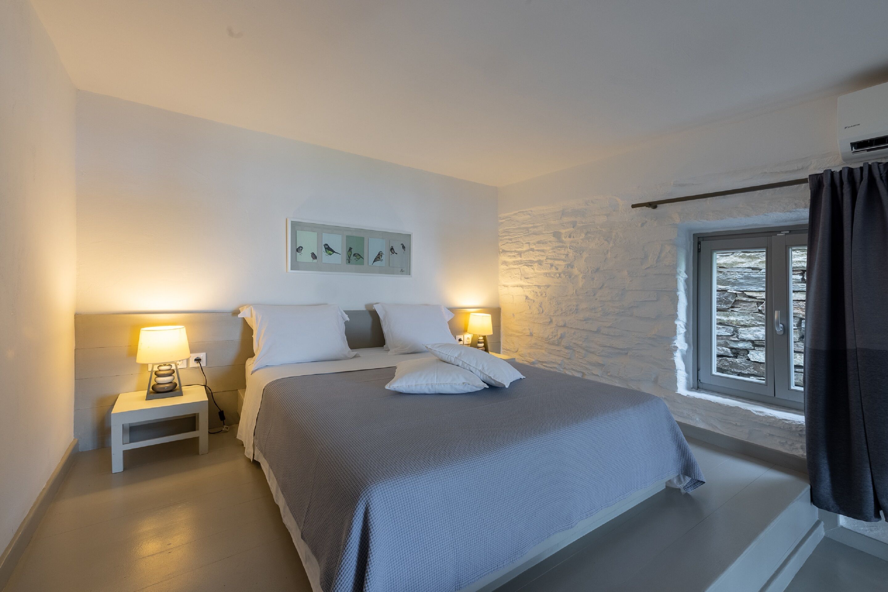 A bedroom with a bed, two bedside tables with lamps, a stone wall, a window with curtains, and a framed picture on the wall.