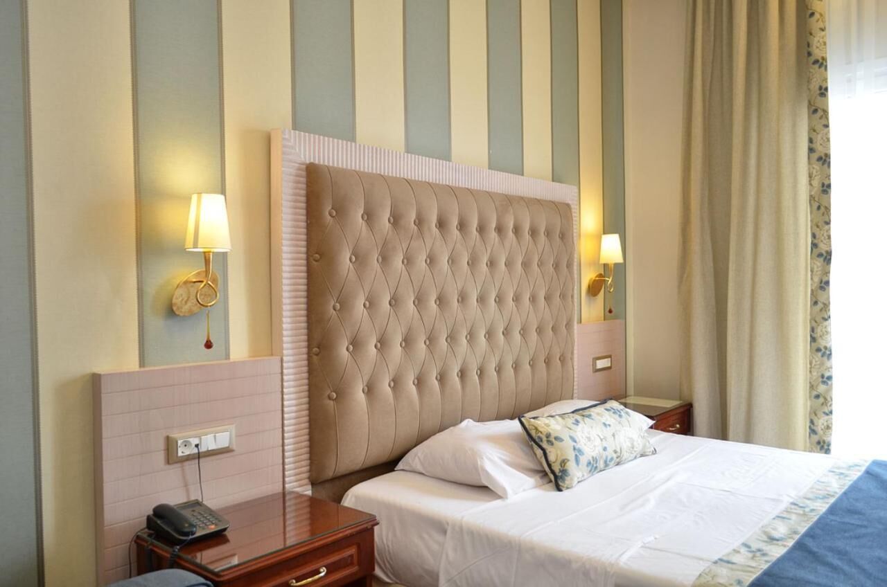 Une chambre d’hôtel avec une tête de lit capitonnée, deux lampes de chevet, un téléphone, une table de chevet et un lit avec du linge de lit blanc et un coussin à motifs floraux.
