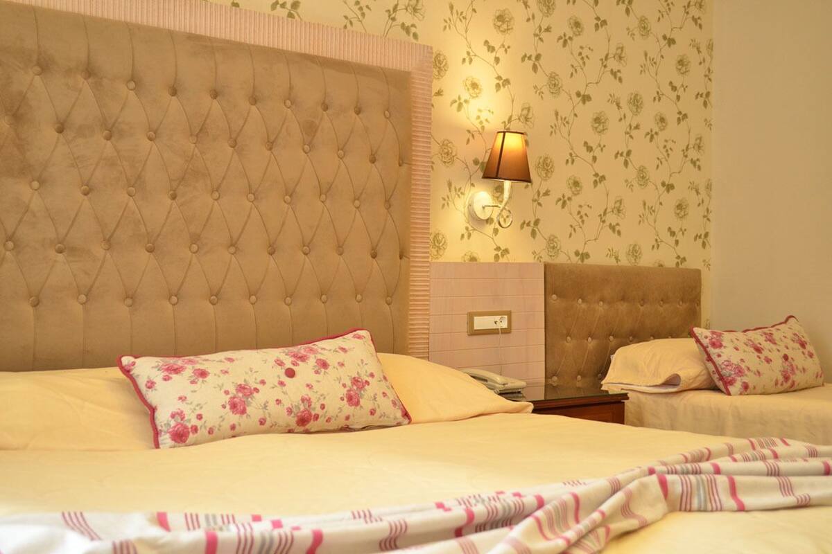 Habitación de hotel con un gran cabecero capitonado, papel tapiz floral, una lámpara de noche y dos camas con almohadas estampadas.