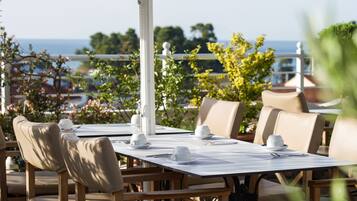 An outdoor dining area with a table set for four, surrounded by beige chairs, overlooking a scenic view of the sea and greenery.