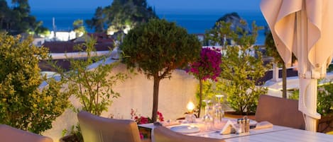 An outdoor dining area with a table set for two, surrounded by chairs, overlooking a scenic view of the sea and distant buildings.