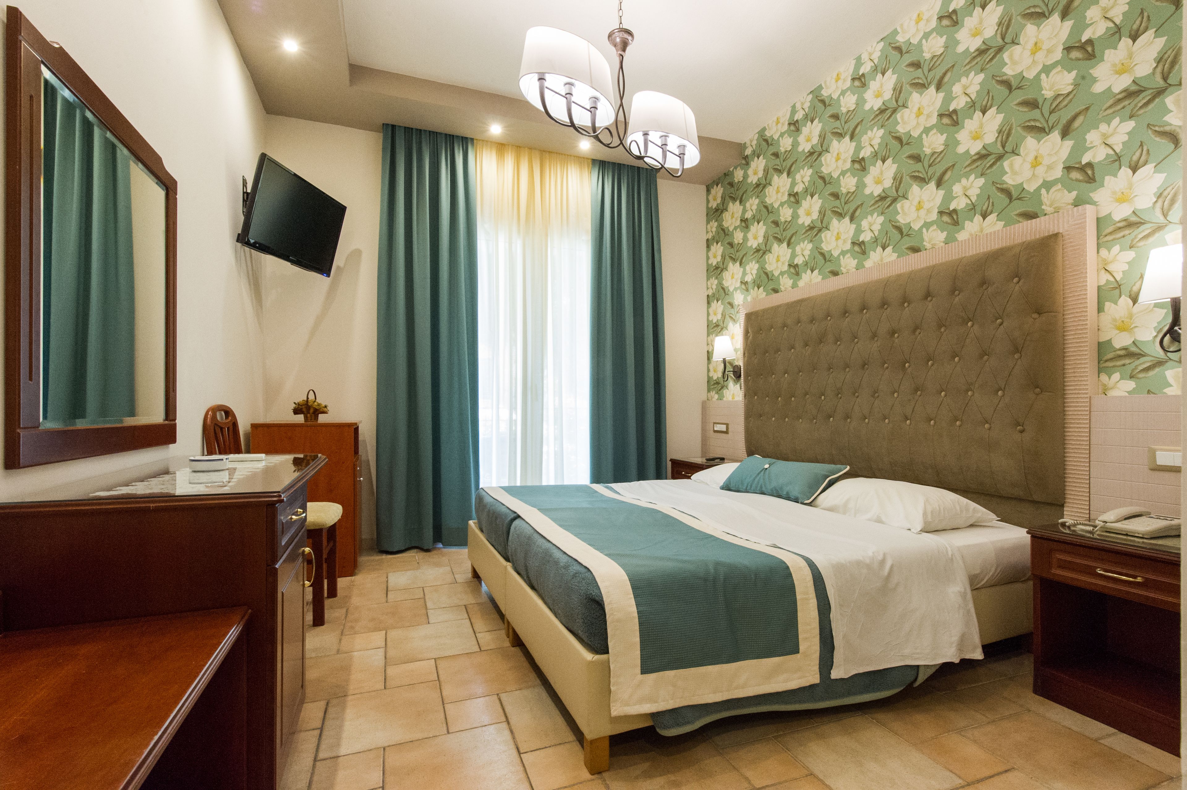 A hotel room with a bed, a desk, a chair, a television, and a wall with floral wallpaper.
