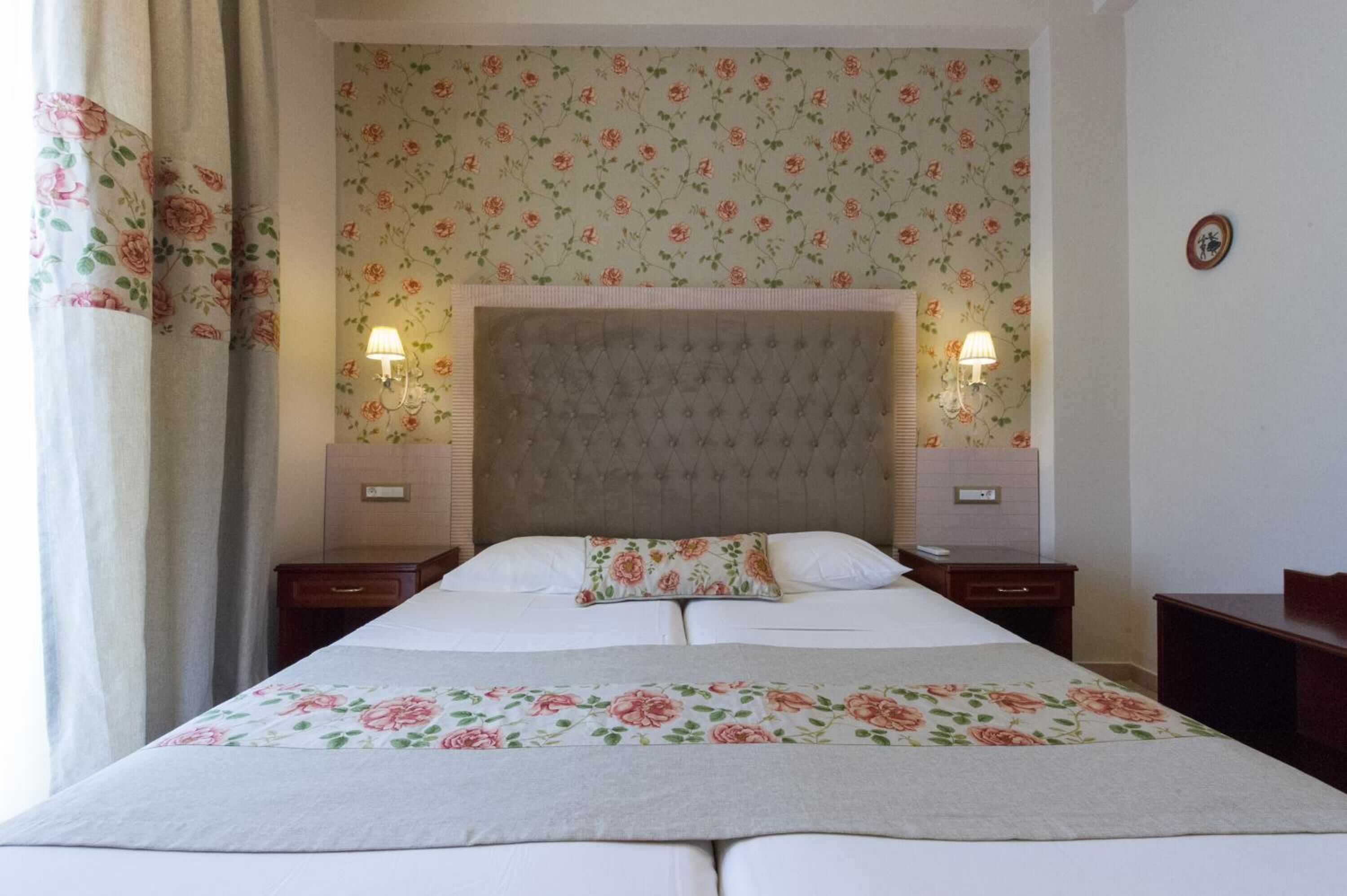 Una habitación de hotel ordenada con una cama grande, papel tapiz floral, dos lámparas de noche y un cabecero.