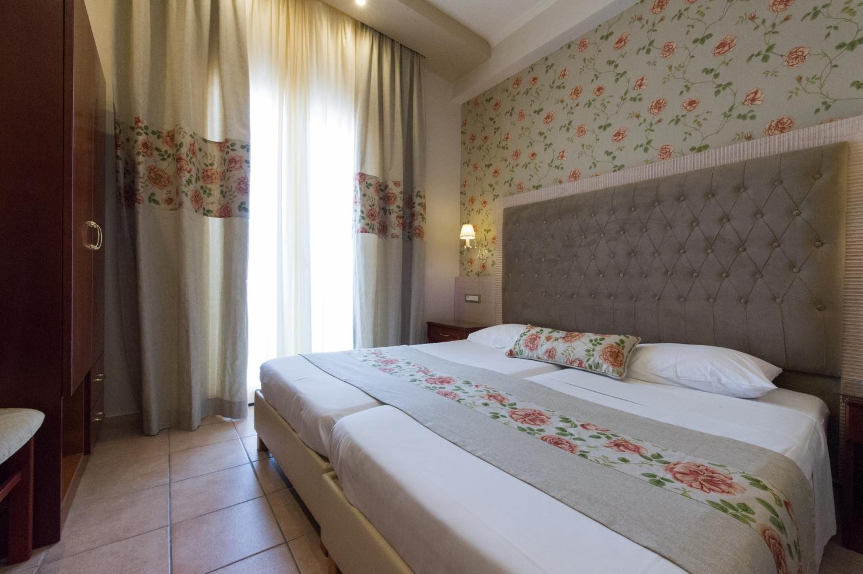A hotel room with a large bed, floral curtains, a tufted headboard, and a floral wallpaper pattern.