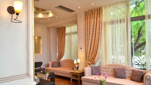 A spacious living area with a sofa, armchairs, and a coffee table. Large windows with curtains provide natural light and a view of greenery outside. - Olympic Hotel (Parga)