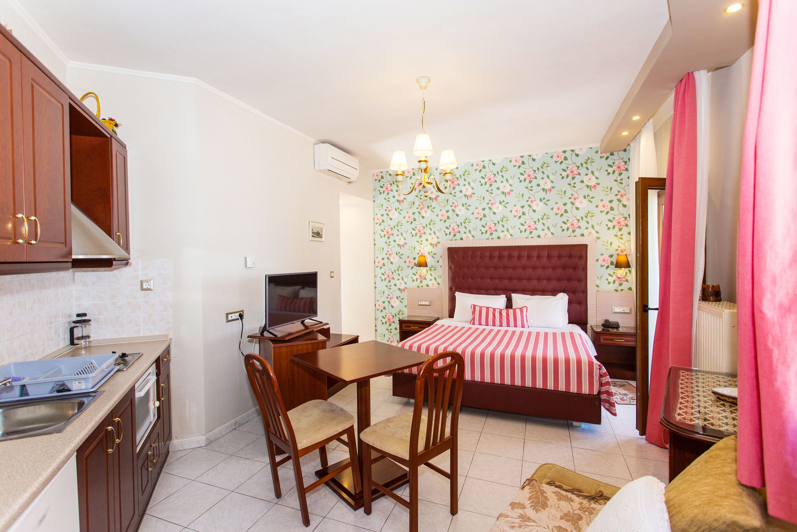 Quarto de hotel compacto com cozinha compacta, cama com cabeceira estampada, área de refeições e um pequeno espaço de estar.