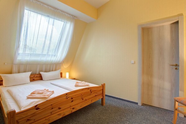 Un dormitorio con cama de madera, ropa de cama blanca, una ventana con cortinas traslúcidas y una puerta.