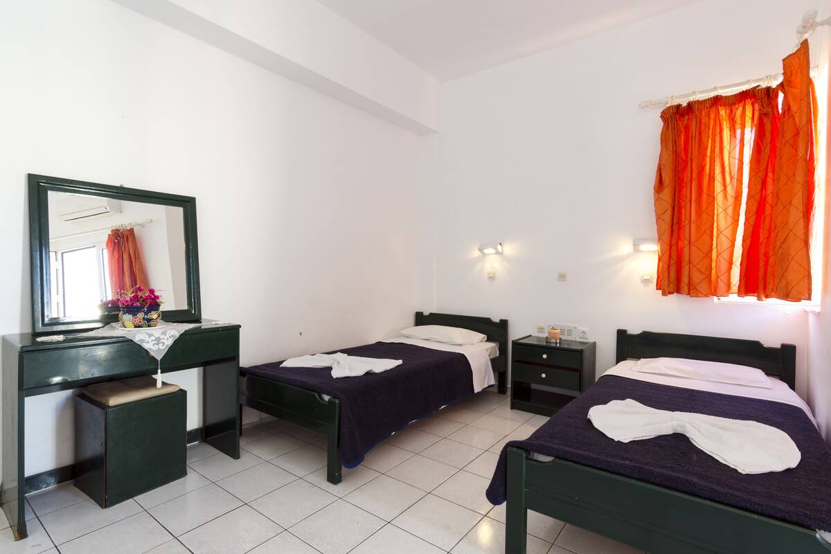 A room with two single beds, a desk with a mirror, a small bedside table, and a window with orange curtains.