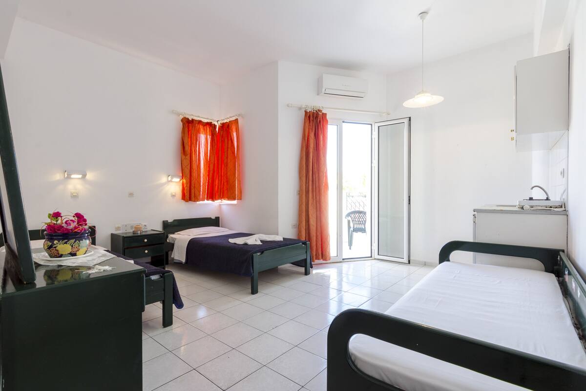 A hotel room with a bed, a desk, a chair, a window with curtains, and a door leading to a balcony.