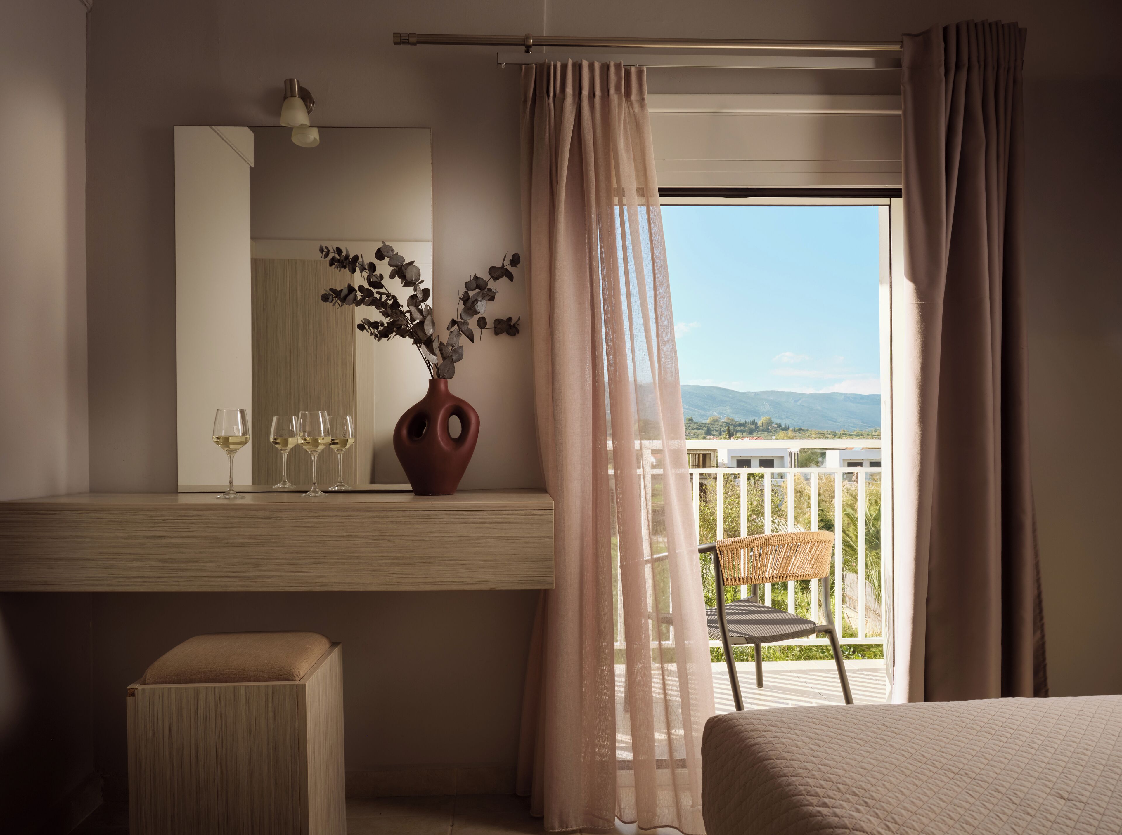 A hotel room with a wooden vanity, a vase with flowers, a chair, and a balcony with a view of mountains.