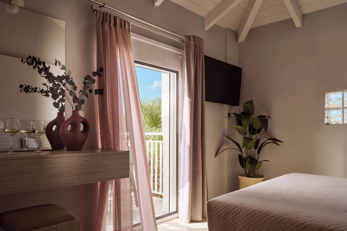A bedroom with a bed, a TV mounted on the wall, a balcony with a view, and a bedside table with a vase and glasses.