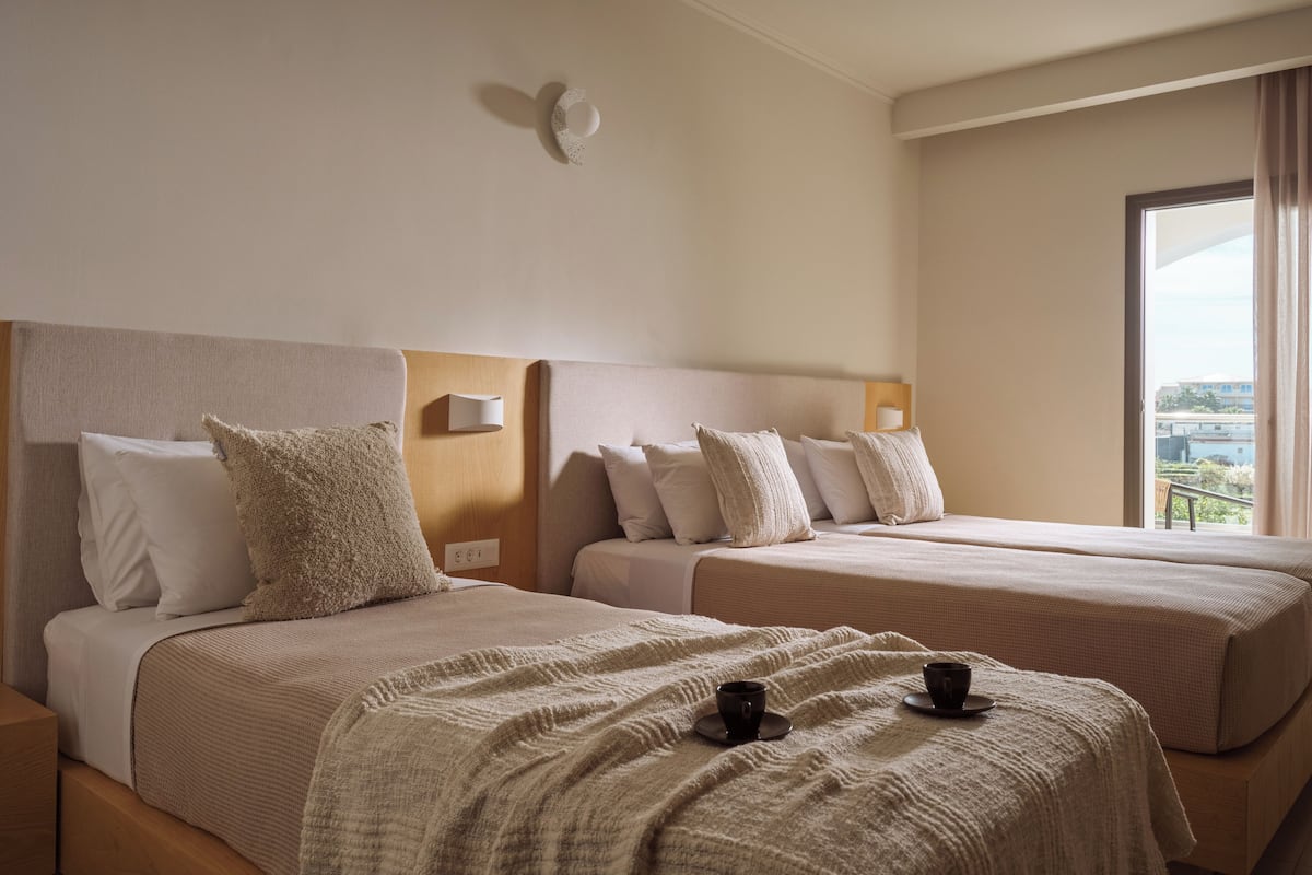 A hotel room with two beds, each with a headboard, pillows, and a blanket. There are two cups on the beds.