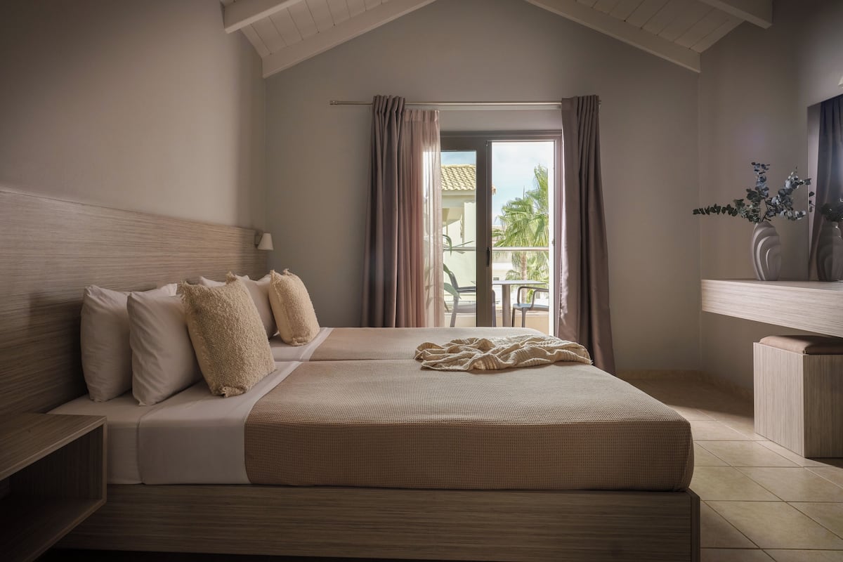 A bedroom with a bed, a nightstand, a lamp, and a view of the outdoors through a window.