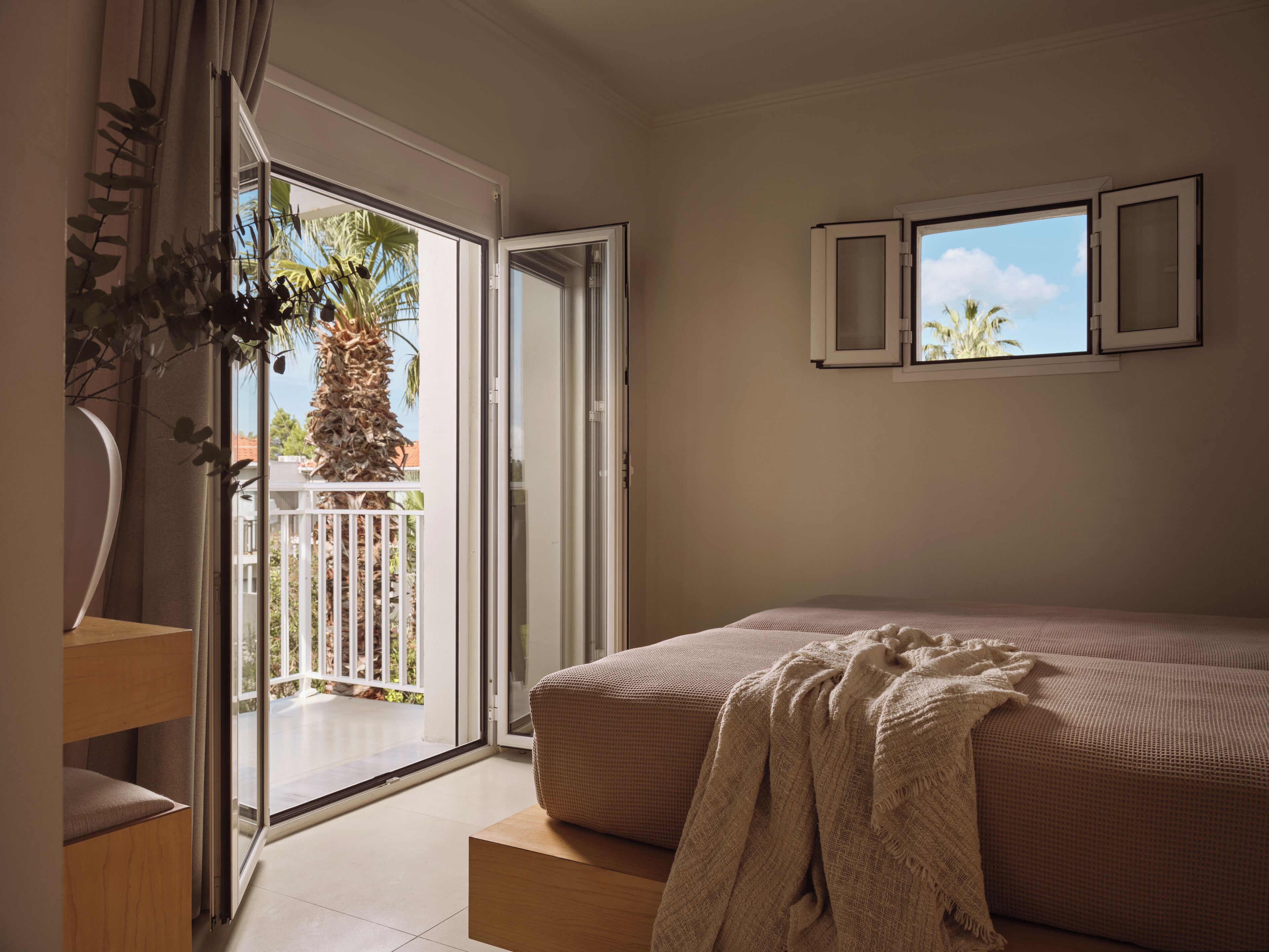 A bedroom with a bed, a balcony with a view of palm trees, and a window with curtains.