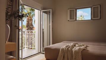 A bedroom with a bed, a balcony with a view of palm trees, and a window with curtains.