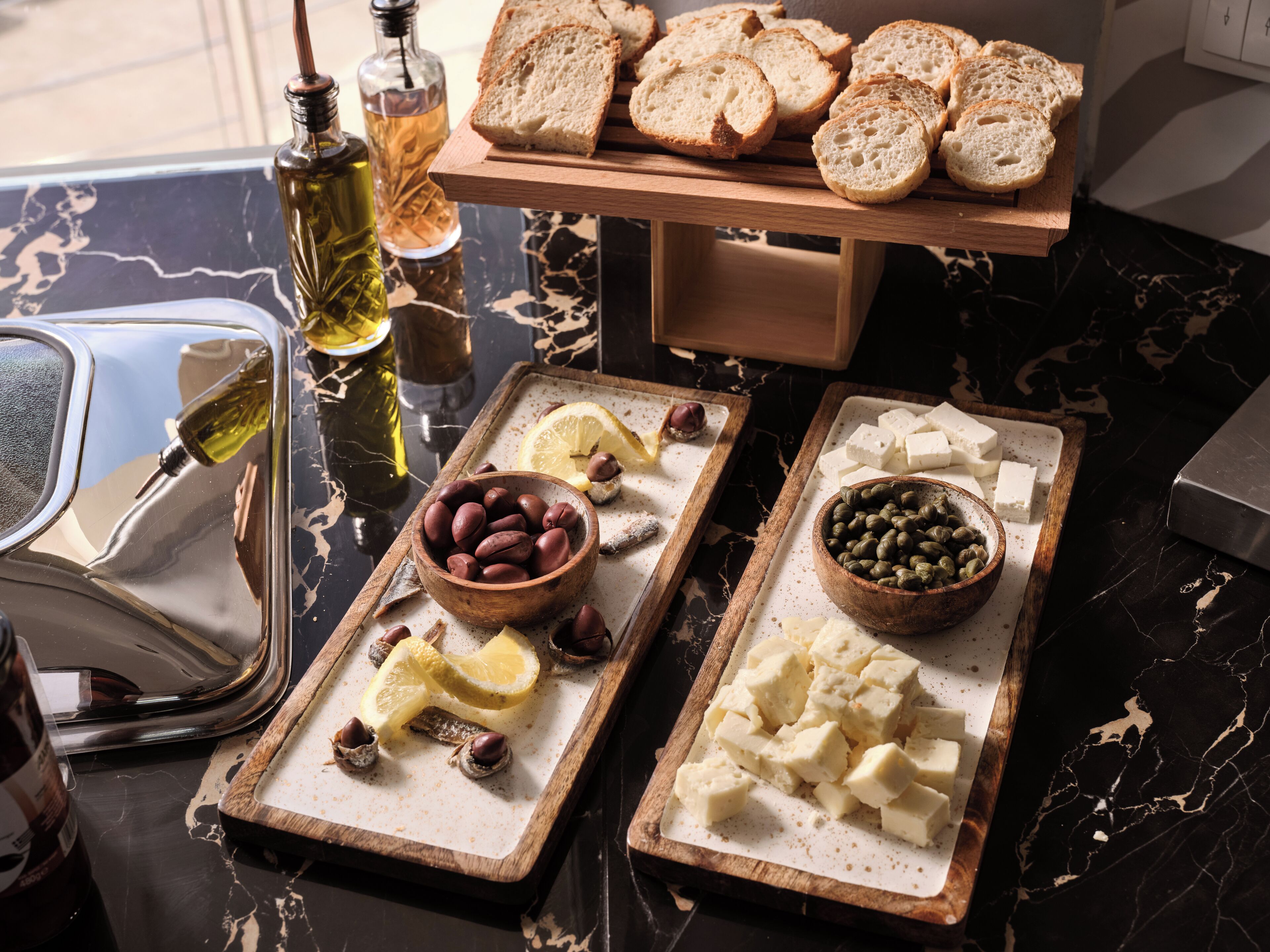 Una encimera de mármol con una bandeja de madera que contiene aceitunas, rodajas de limón y frutos secos; una tabla de madera con rebanadas de pan; y una mesa de mármol con una botella de aceite de oliva y una copa.