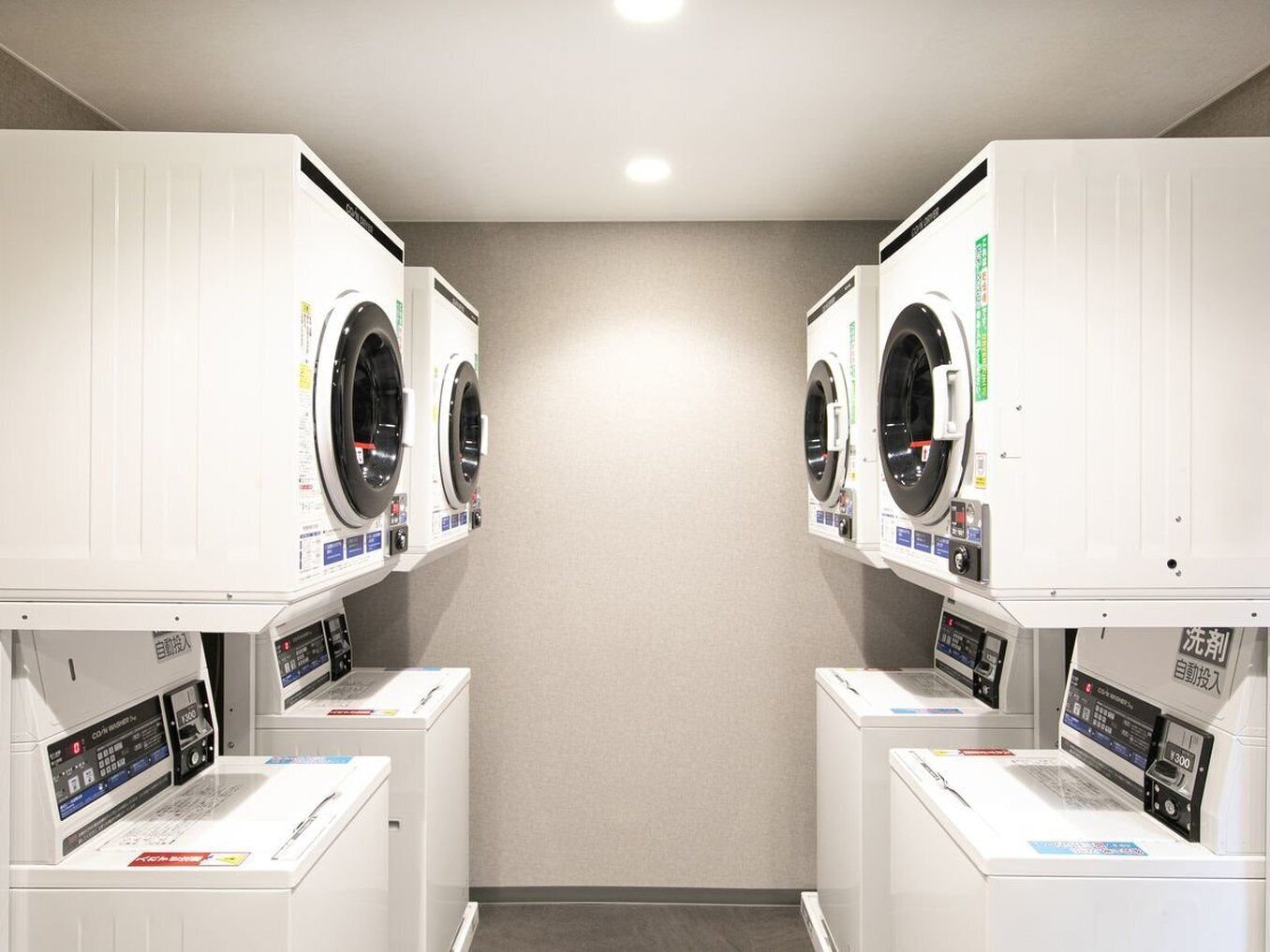 A laundry facility with two rows of washing machines and dryers, a control panel, and a grey wall.
