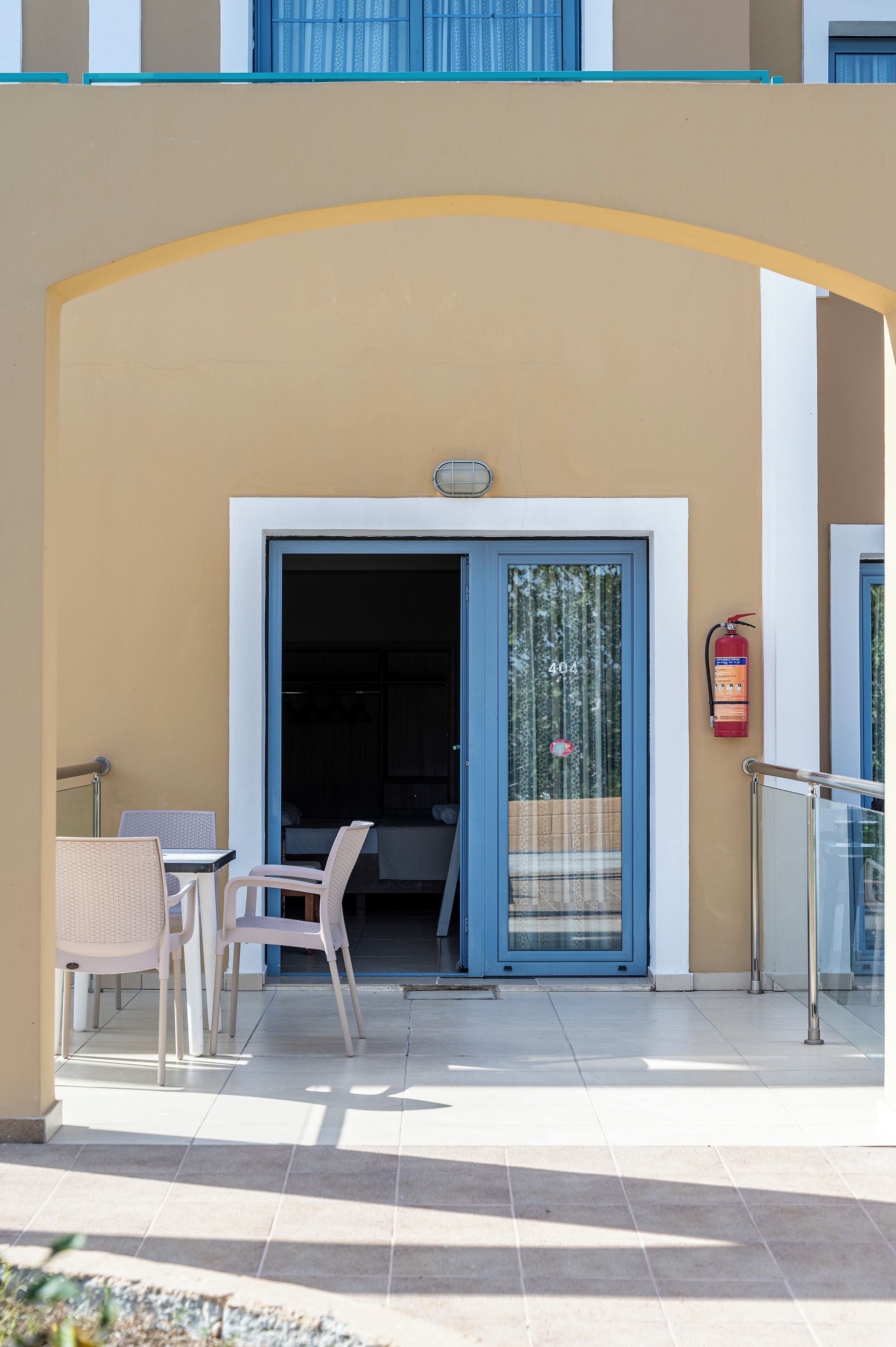 A hotel entrance with a glass door, a fire extinguisher mounted on the wall, and a small outdoor seating area with chairs.