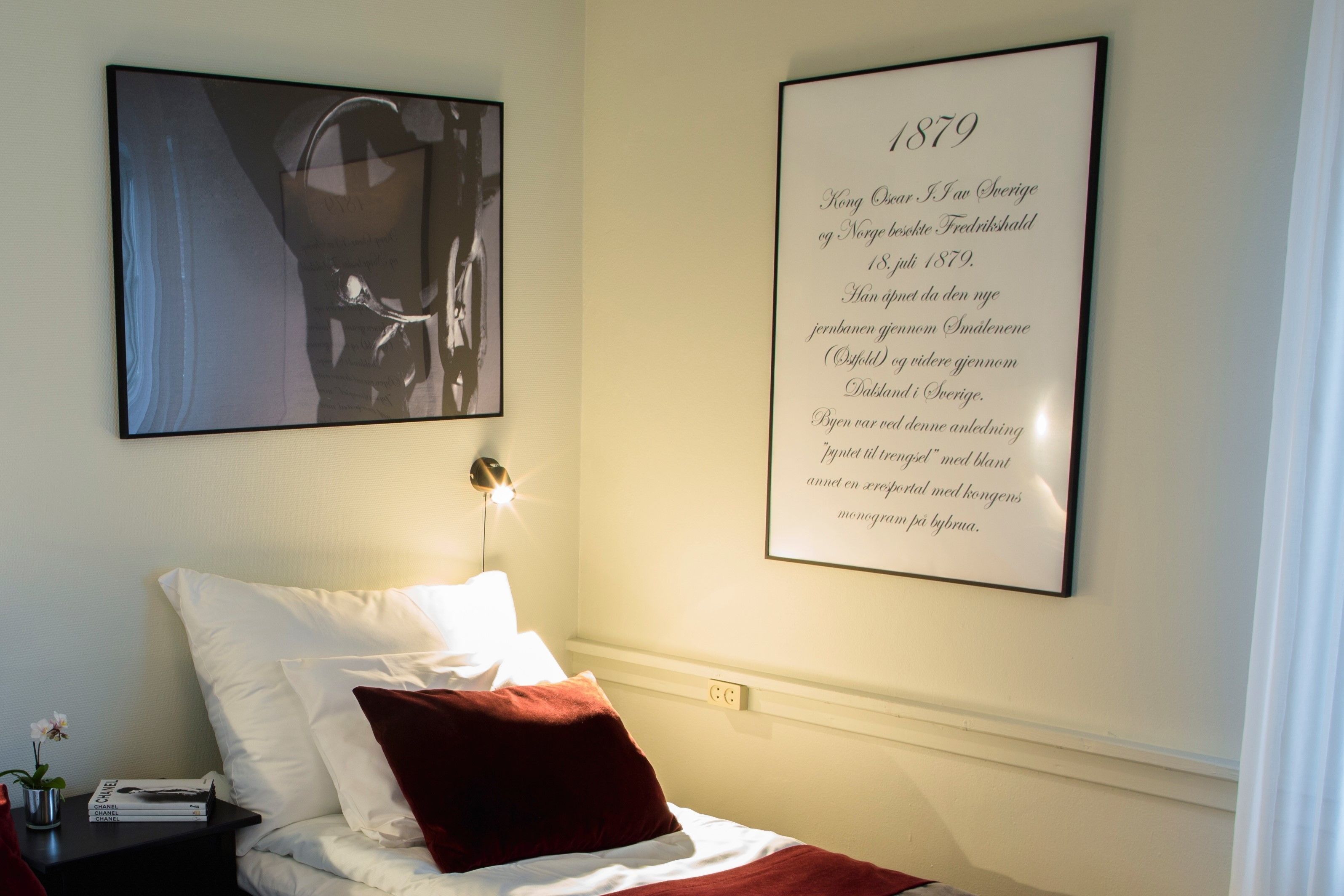 A bedroom with a bed, pillows, a nightstand with books and a vase, a framed text on the wall, and a framed photo above the bed.