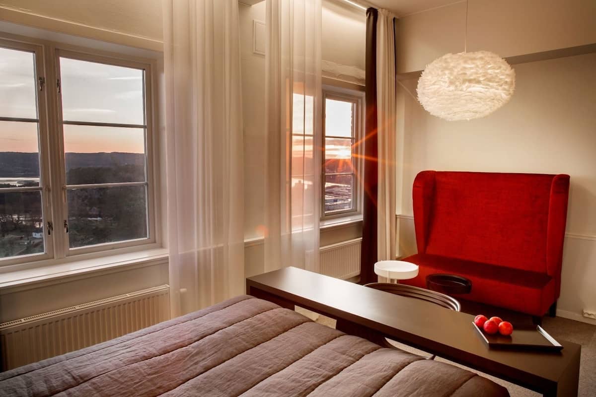 A hotel room with a bed, a red chair, a table with fruit, and a view of the city through large windows.