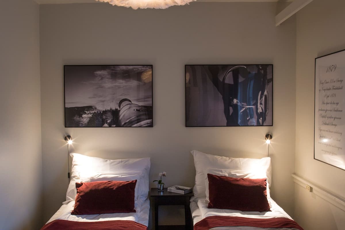 A hotel room with two single beds, each with a red pillow and white headboard. There is a large, fluffy ceiling light fixture above the beds. Two framed artworks are hung on the wall between the beds.
