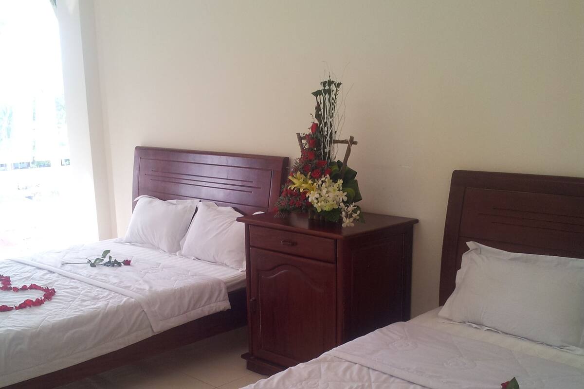 Two double beds with white linens, a wooden nightstand with a vase of flowers, and a heart-shaped arrangement of rose petals on the beds.