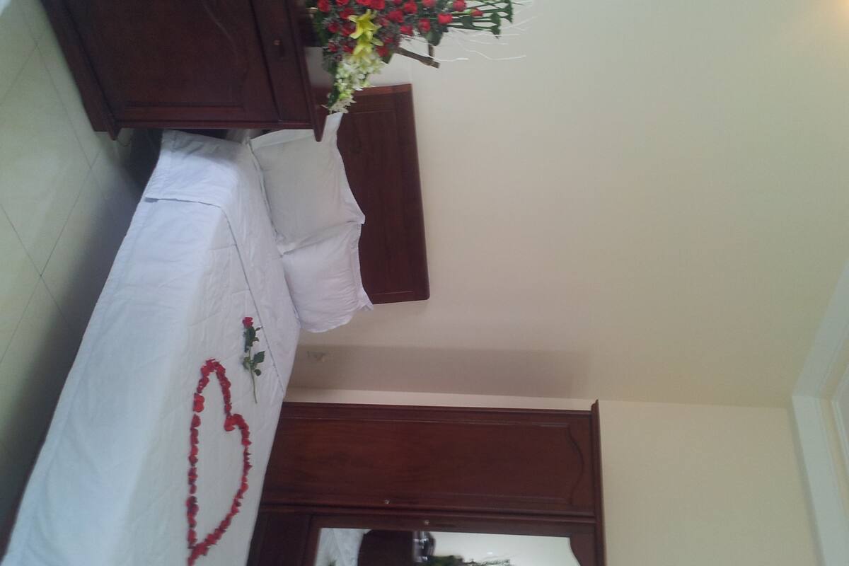 A bedroom with a bed, a wooden wardrobe, and a floral arrangement on the nightstand.