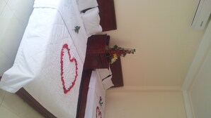 A neatly arranged bed with a heart-shaped decoration on the bedspread, a wooden nightstand with a vase of flowers, and a white wall with a thermostat. - Seaside Hotel 2 (Nha Trang)