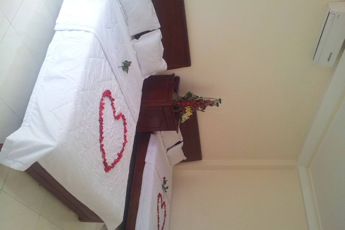 A neatly arranged bed with a heart-shaped decoration on the bedspread, a wooden nightstand with a vase of flowers, and a white wall with a thermostat.