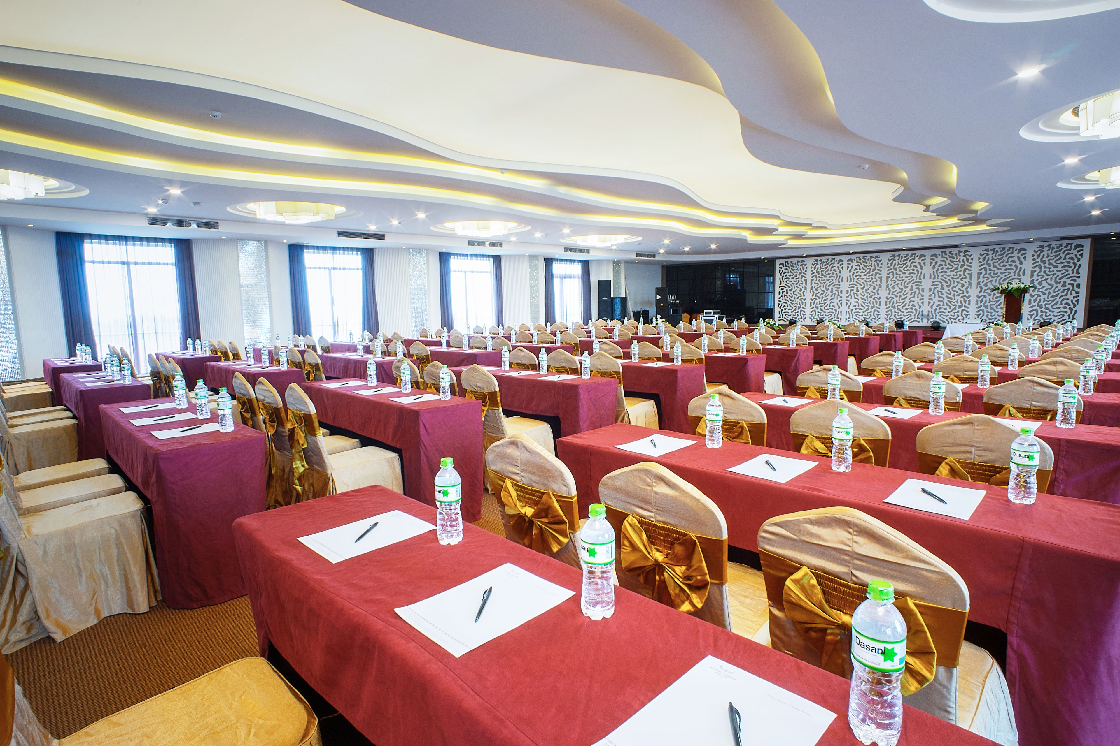 Une salle de conférence avec des rangées de tables, chacune recouverte d’une nappe rouge, et sur laquelle on trouve du papier à lettres blanc, des stylos et des bouteilles d’eau.