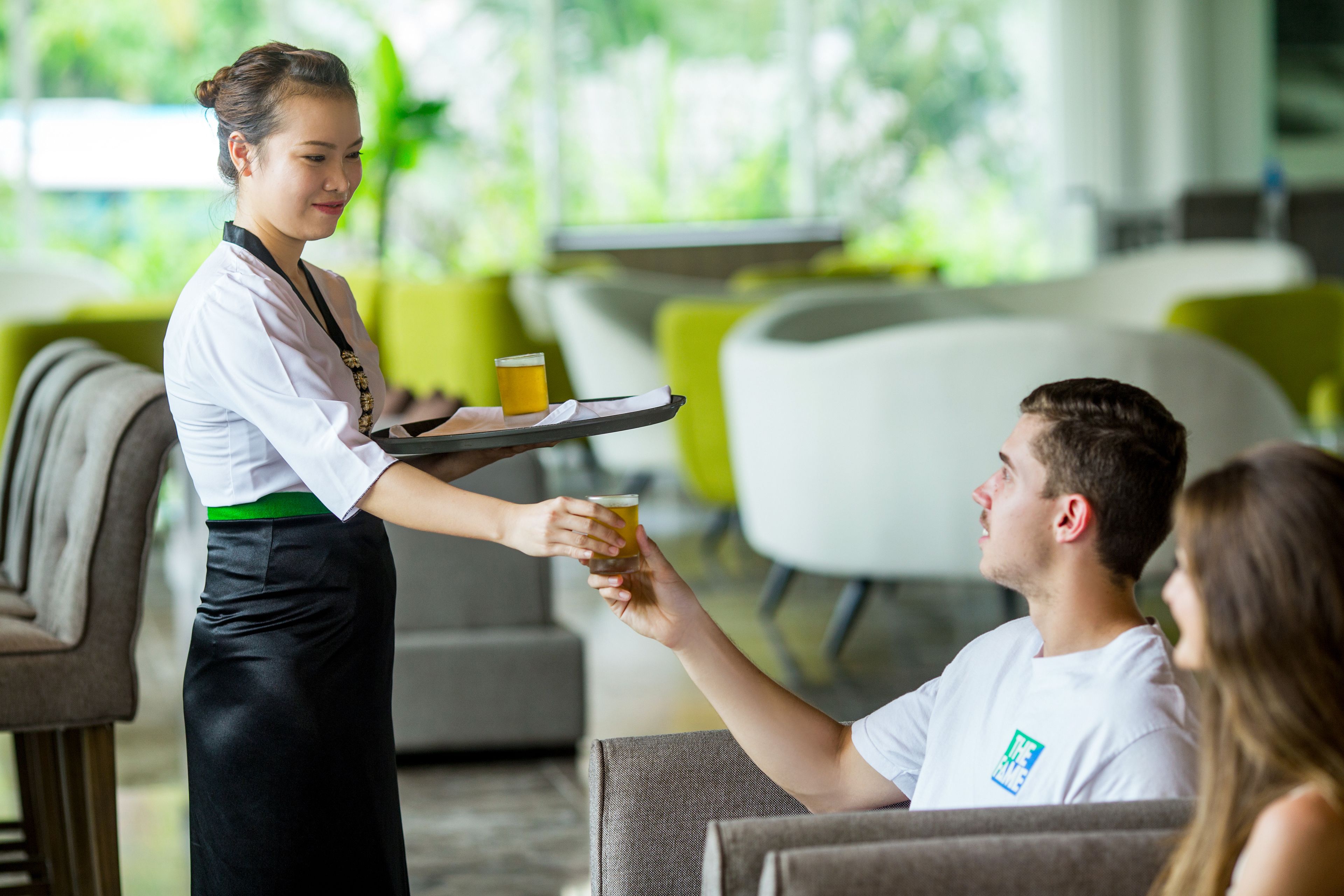 Une personne tenant un plateau avec une boisson s’approche d’une autre personne assise dans un espace intérieur moderne et bien éclairé, aménagé avec des meubles contemporains.