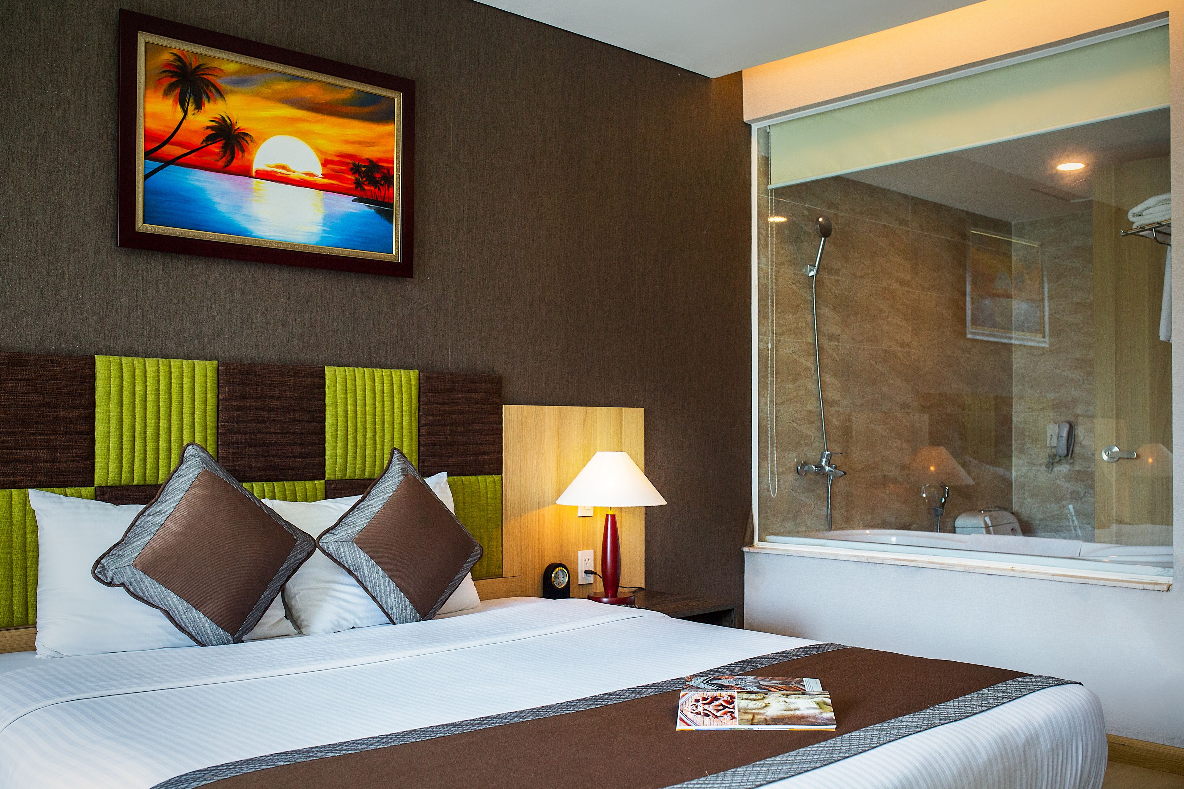 Habitación de hotel con cama, mesita de noche, lámpara, un cuadro de un atardecer y zona de ducha.